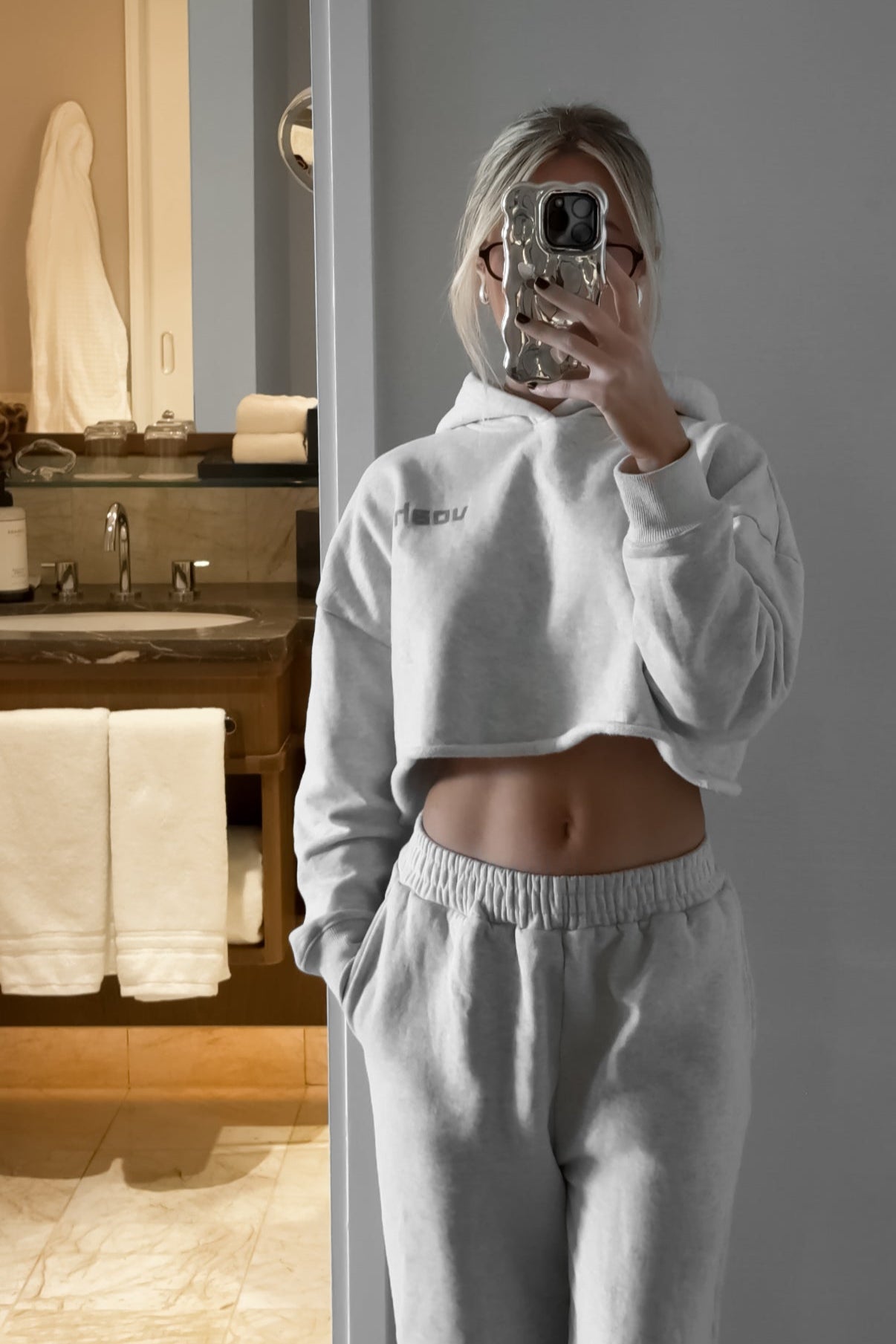 Person taking a mirror selfie wearing a gray hoodie and pants in a bathroom.